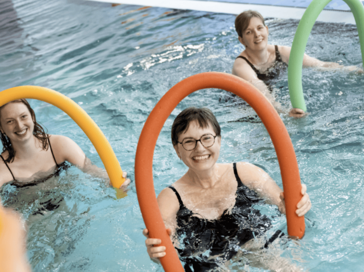Lachende Frauen im Aquafitness-Kurs mit Schwimmnudeln