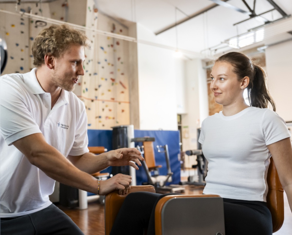 Ein junger Mitarbeiter des medicos.Osnabrück zeigt einer Patientin die korrekte Nutzung eines Fitnessgeräts – individuelle Betreuung und therapeutisch begleitetes Training in angenehmer Atmosphäre